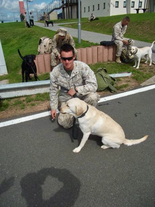 Soldier with dog outdoors, others in background.